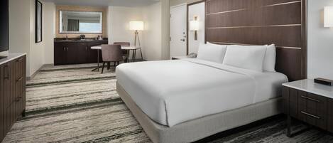 A hotel room with a large bed, a desk, a chair, a desk lamp, a mirror, and a bathroom with a sink and mirror.