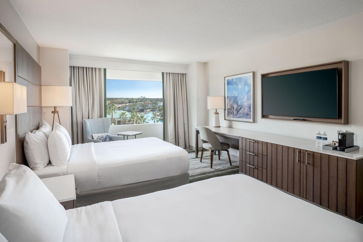 A hotel room with two beds, a desk, a TV, and a view of a resort.
