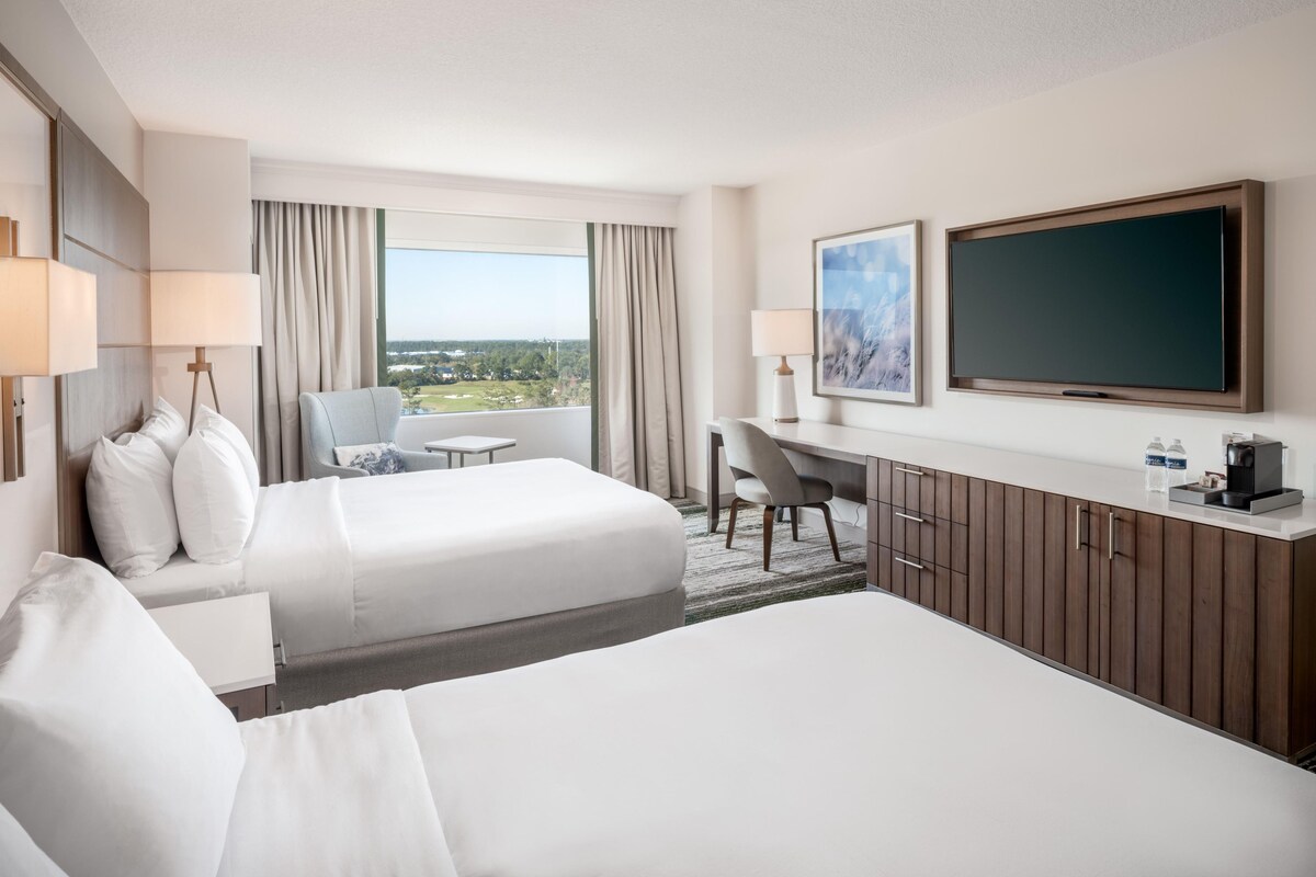 A hotel room with two beds, a desk, a chair, a TV, and a window with a view.