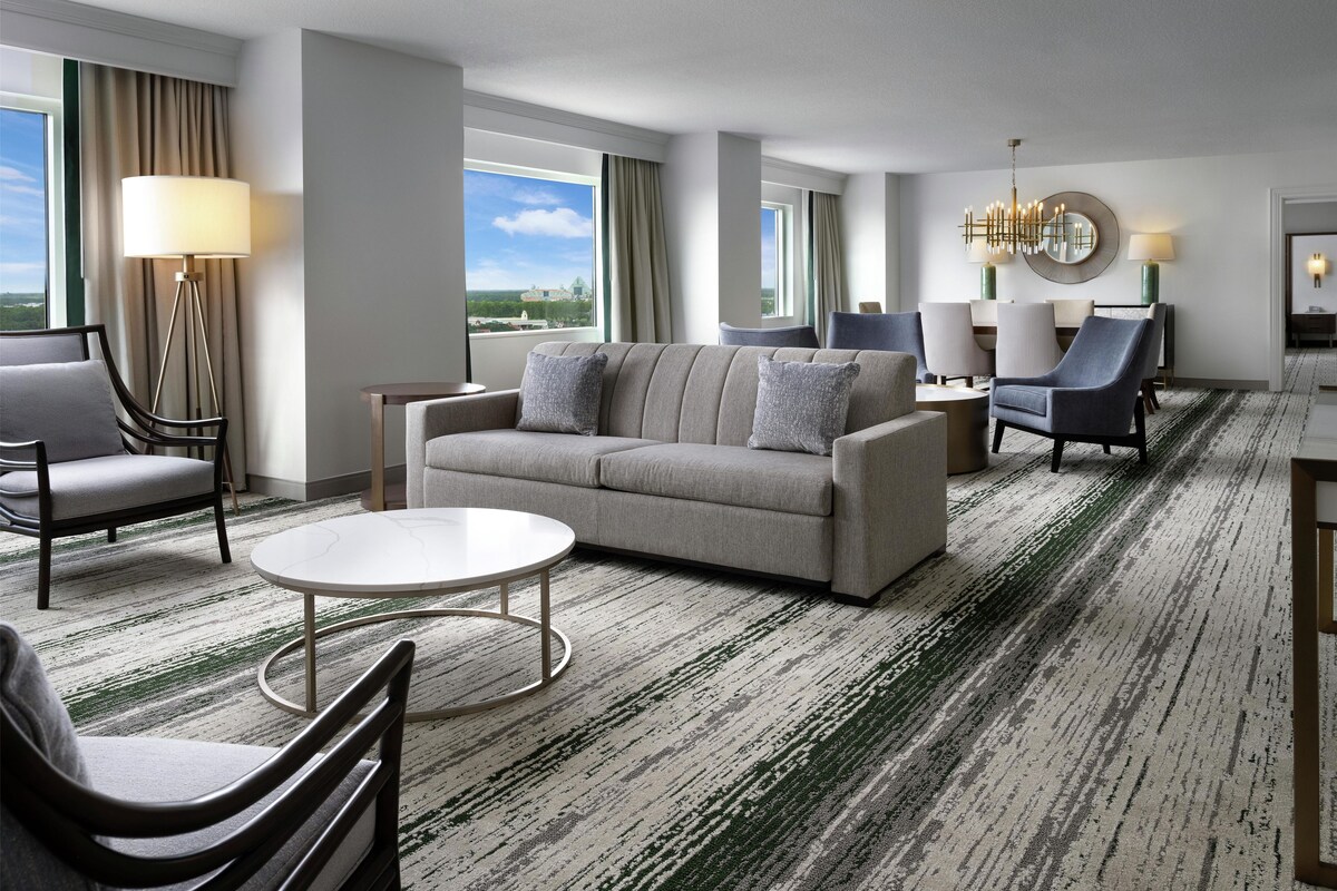 A hotel room with a sofa, armchairs, a coffee table, a dining table with chairs, a chandelier, and large windows with curtains.