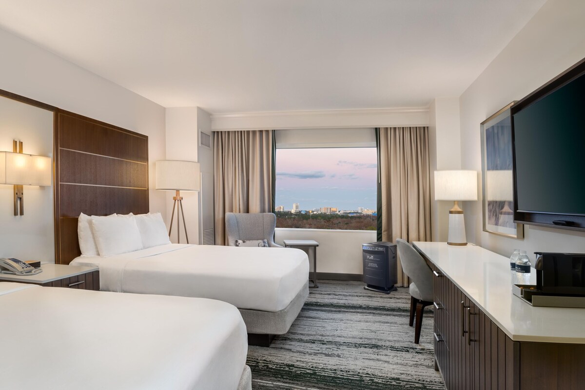 A hotel room with two beds, a desk, a chair, a TV, and a window with a city view.