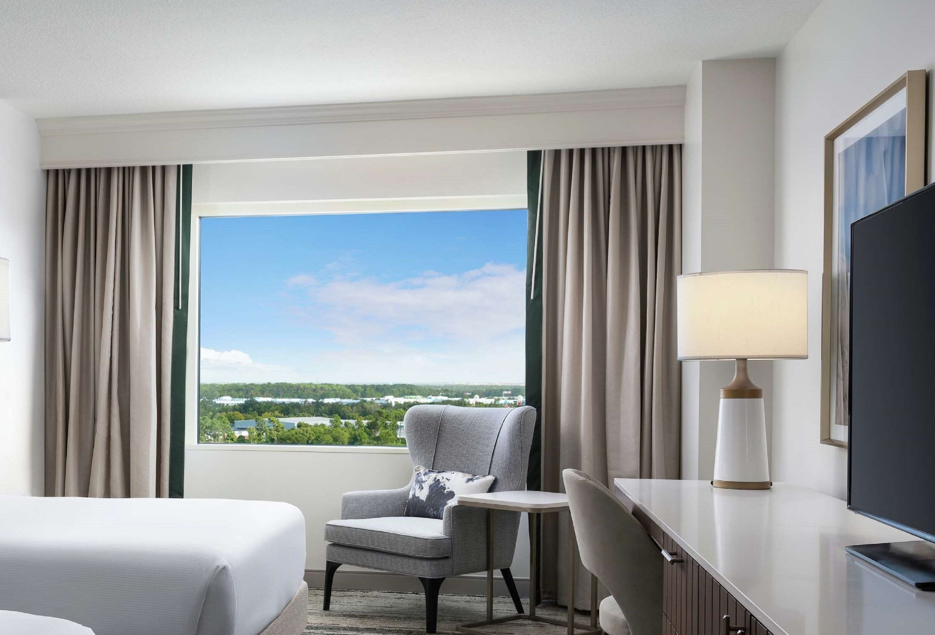 A hotel room with a large window, a bed, a chair, a desk, a lamp, and a television.