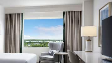 A hotel room with a large window, a bed, a chair, a desk, a lamp, and a television.