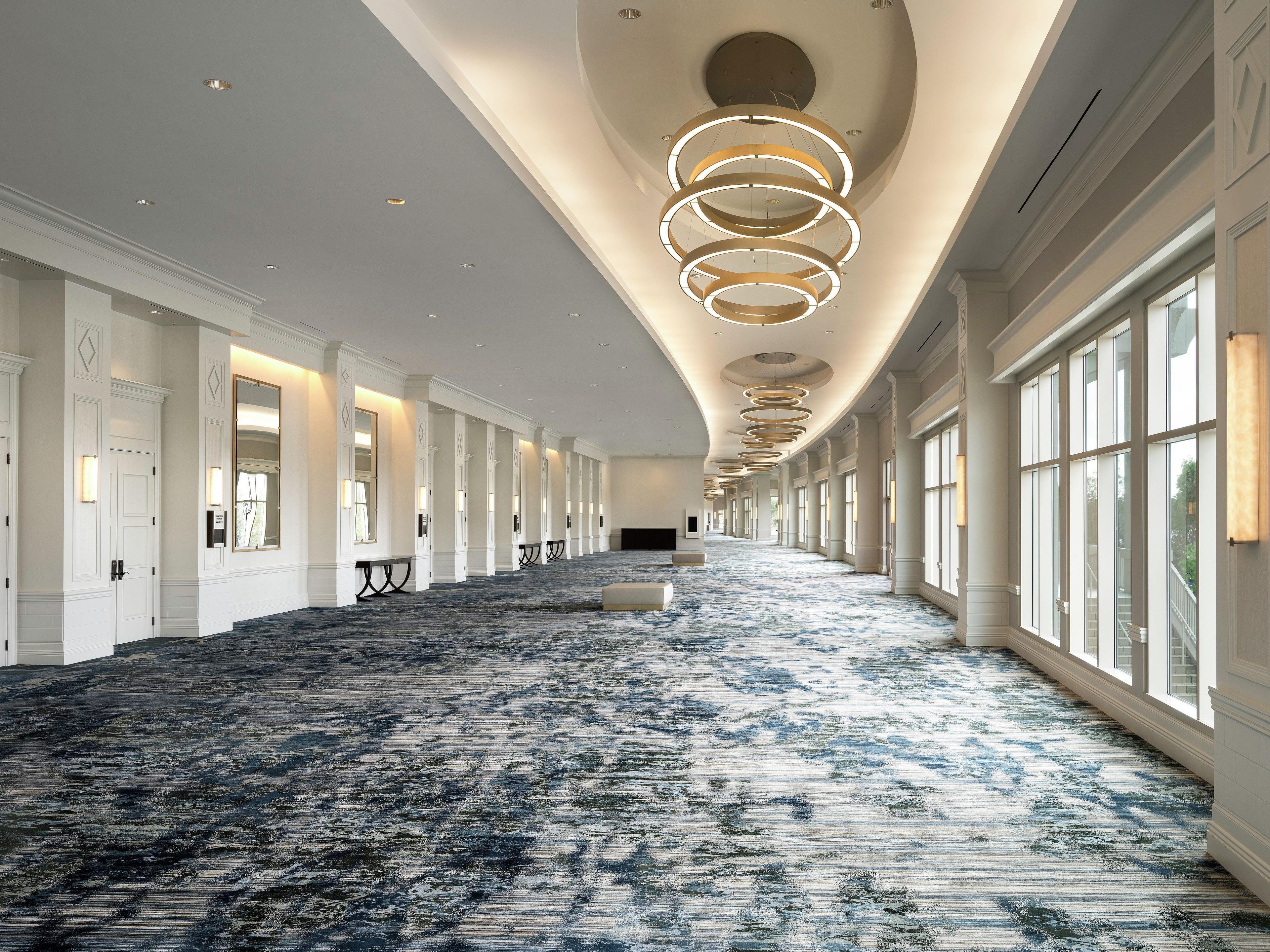 A long corridor with a patterned carpet, large windows, and a series of circular light fixtures on the ceiling.