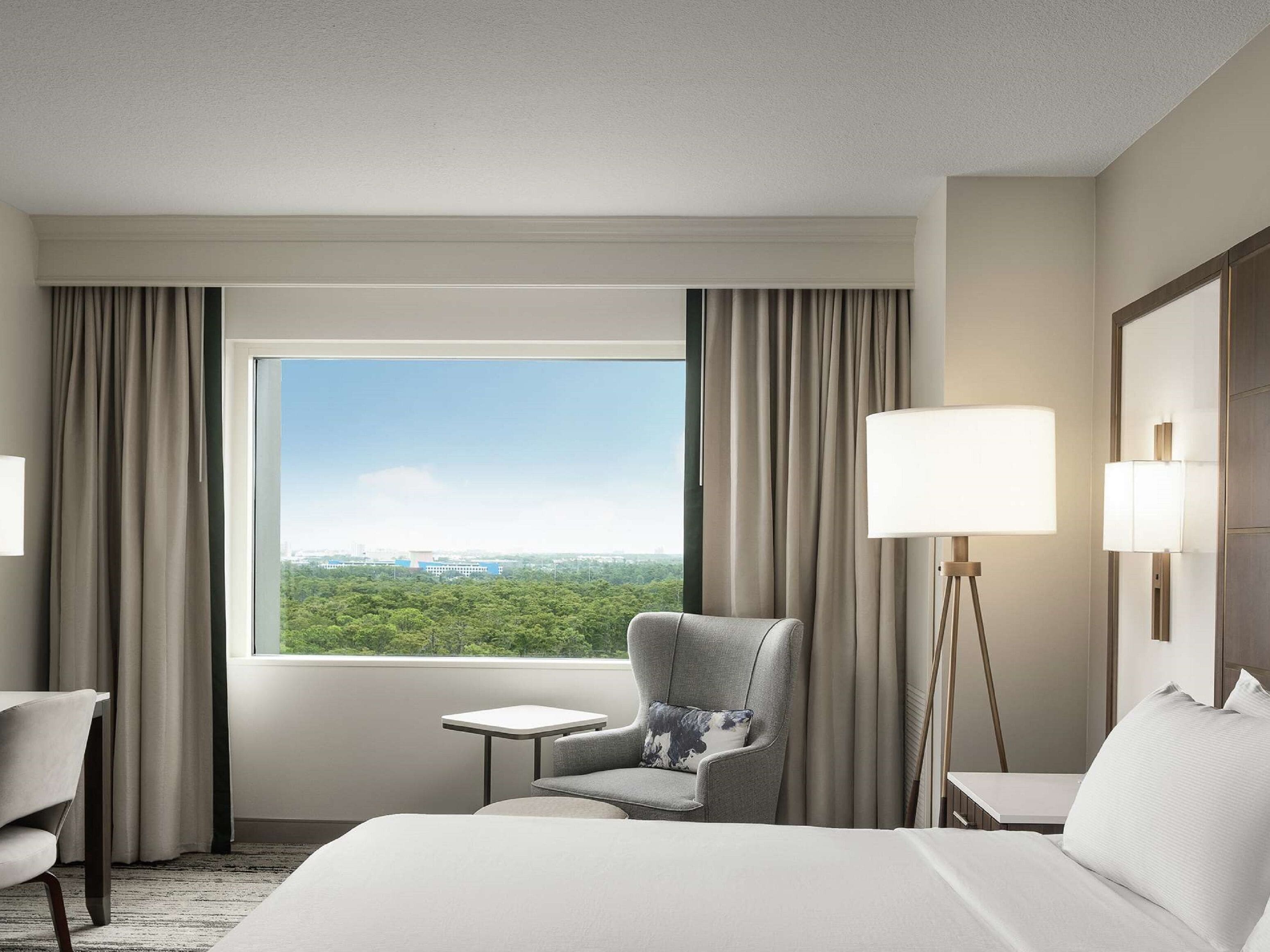 Habitación de hotel con una cama grande, una silla, una mesita y una lámpara. Se ve una ventana con vistas a zonas verdes y edificios.