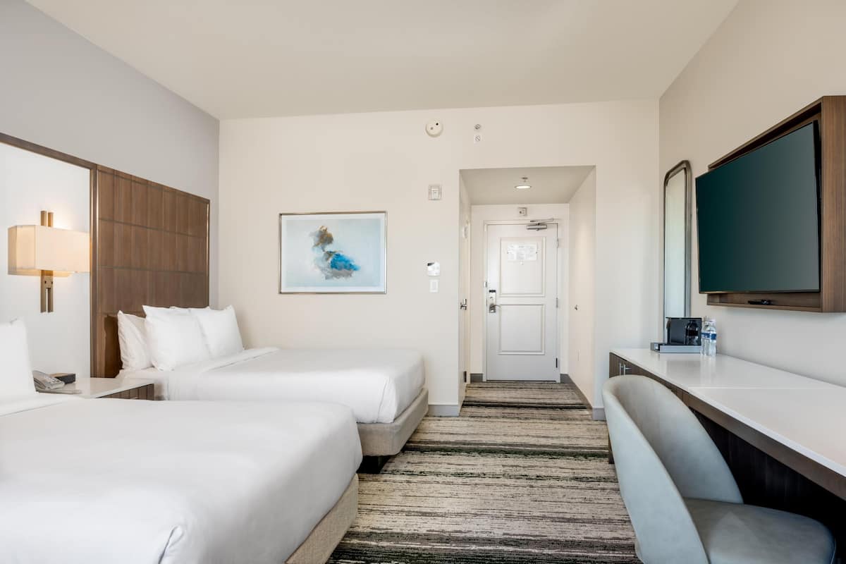Ein Hotelzimmer mit zwei Betten, einem Schreibtisch, einem Sessel, einem Fernseher und einem Gemälde an der Wand.