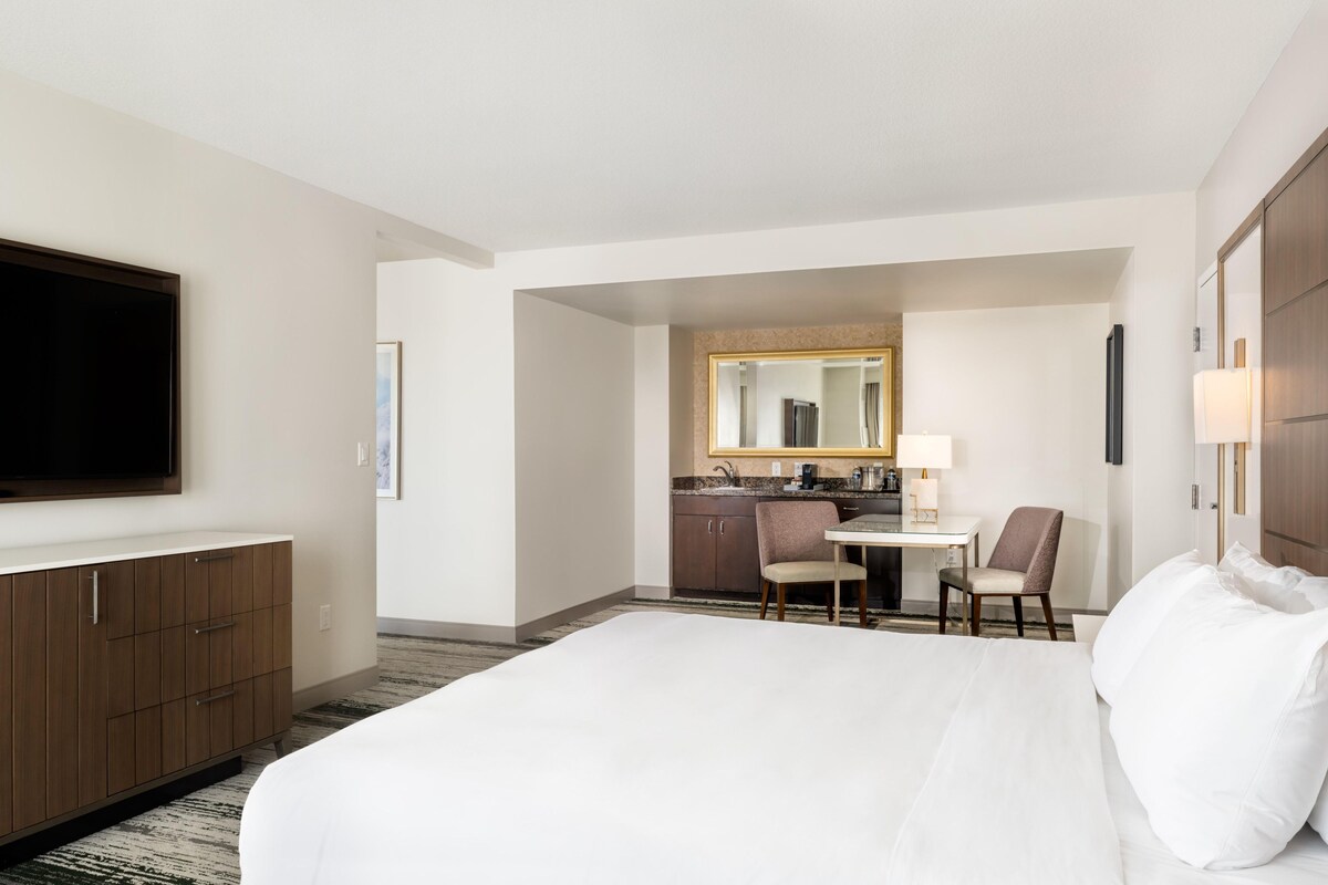 A hotel room with a large bed, a TV, a desk with a chair, and a kitchenette.