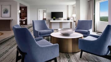 A modern hotel room with a round coffee table, four blue armchairs, a flat-screen TV, and a large window offering a city view.