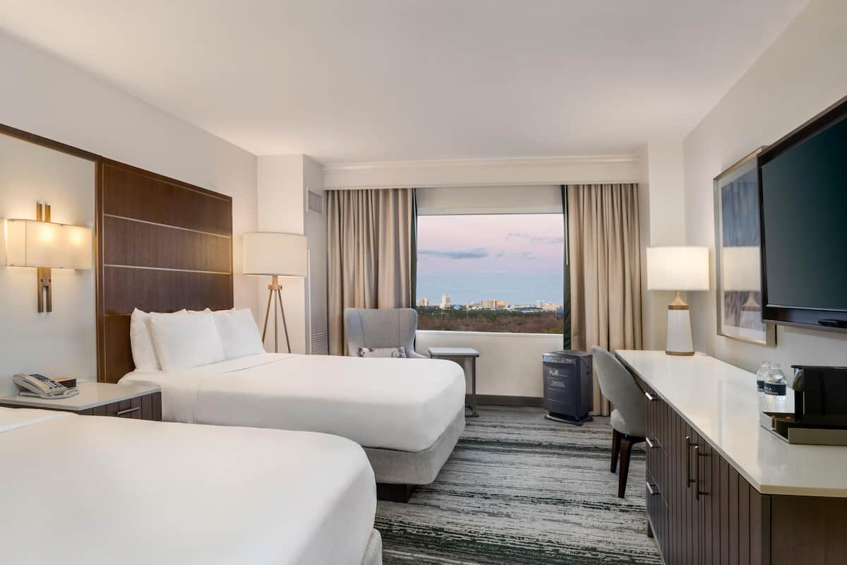 Ein Hotelzimmer mit zwei Betten, einem Schreibtisch, einem Sessel, einem Fernseher und einem Fenster mit Blick auf die Stadt.