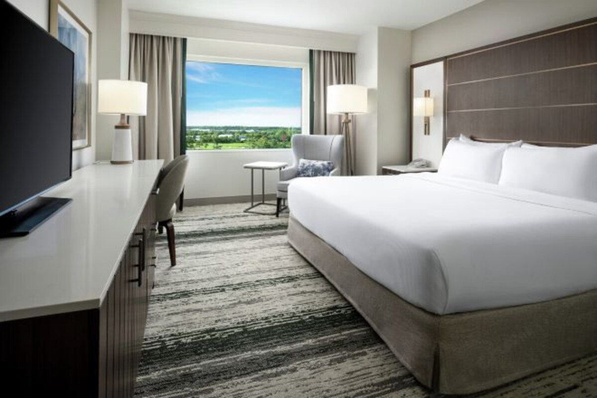 A hotel room with a large bed, a desk with a TV, a chair, a small table, and a view of the outdoors.