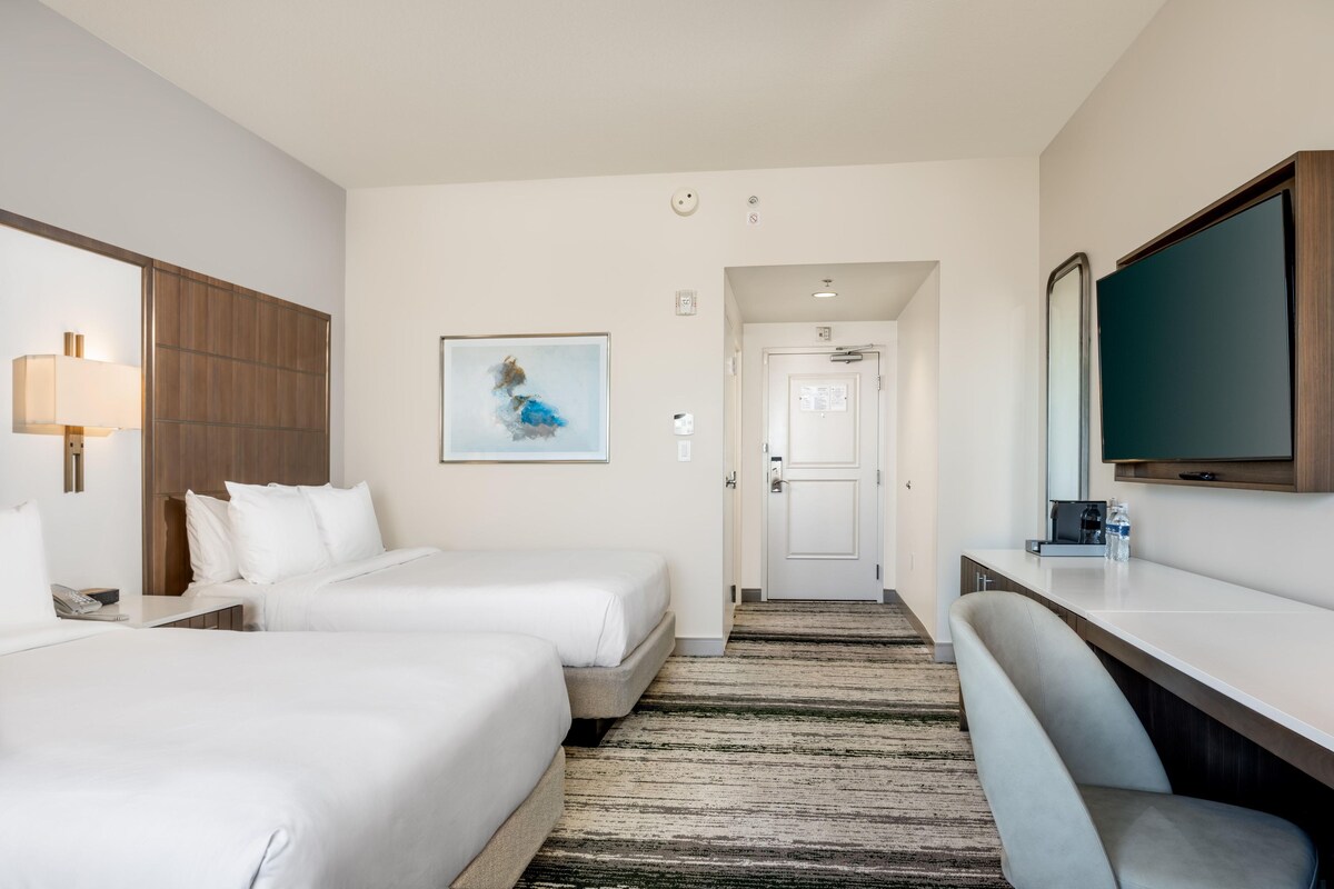 A hotel room with two beds, a desk, a chair, a television, and a painting on the wall.