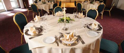 Un salón de banquetes con mesas redondas dispuestas para un evento formal, con sillas doradas y manteles blancos.
