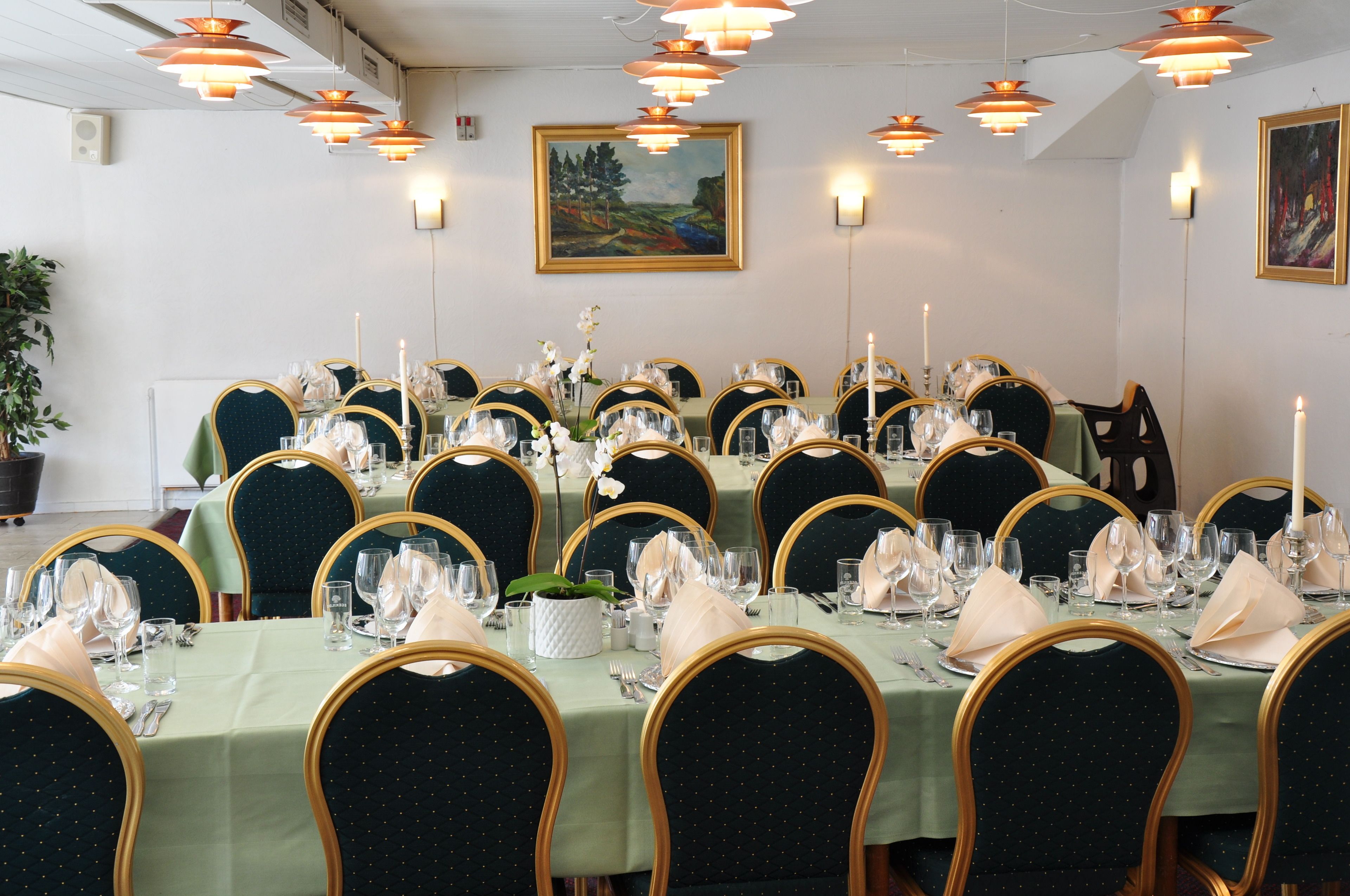 A dining area with round tables set for a meal, featuring green tablecloths, gold chairs, and candle centerpieces.