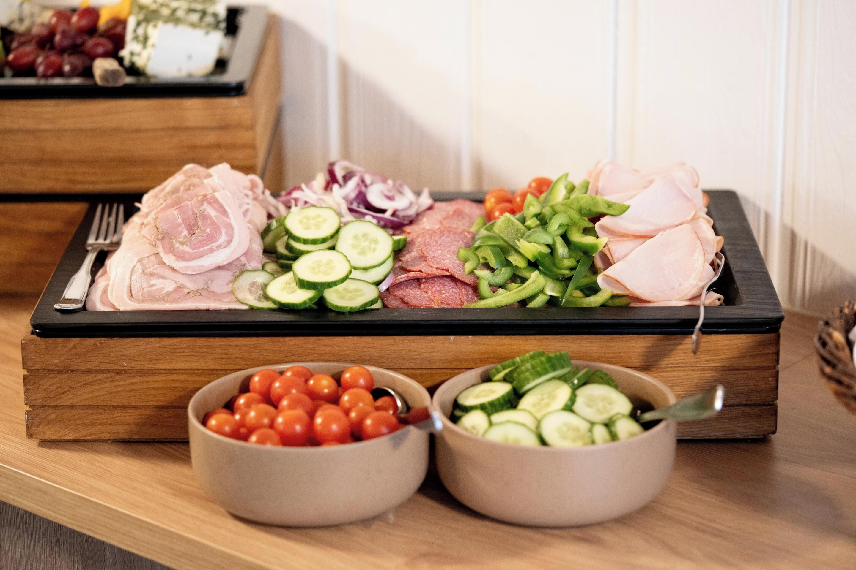 A wooden tray with sliced meats, vegetables, and two bowls of cherry tomatoes and cucumbers.