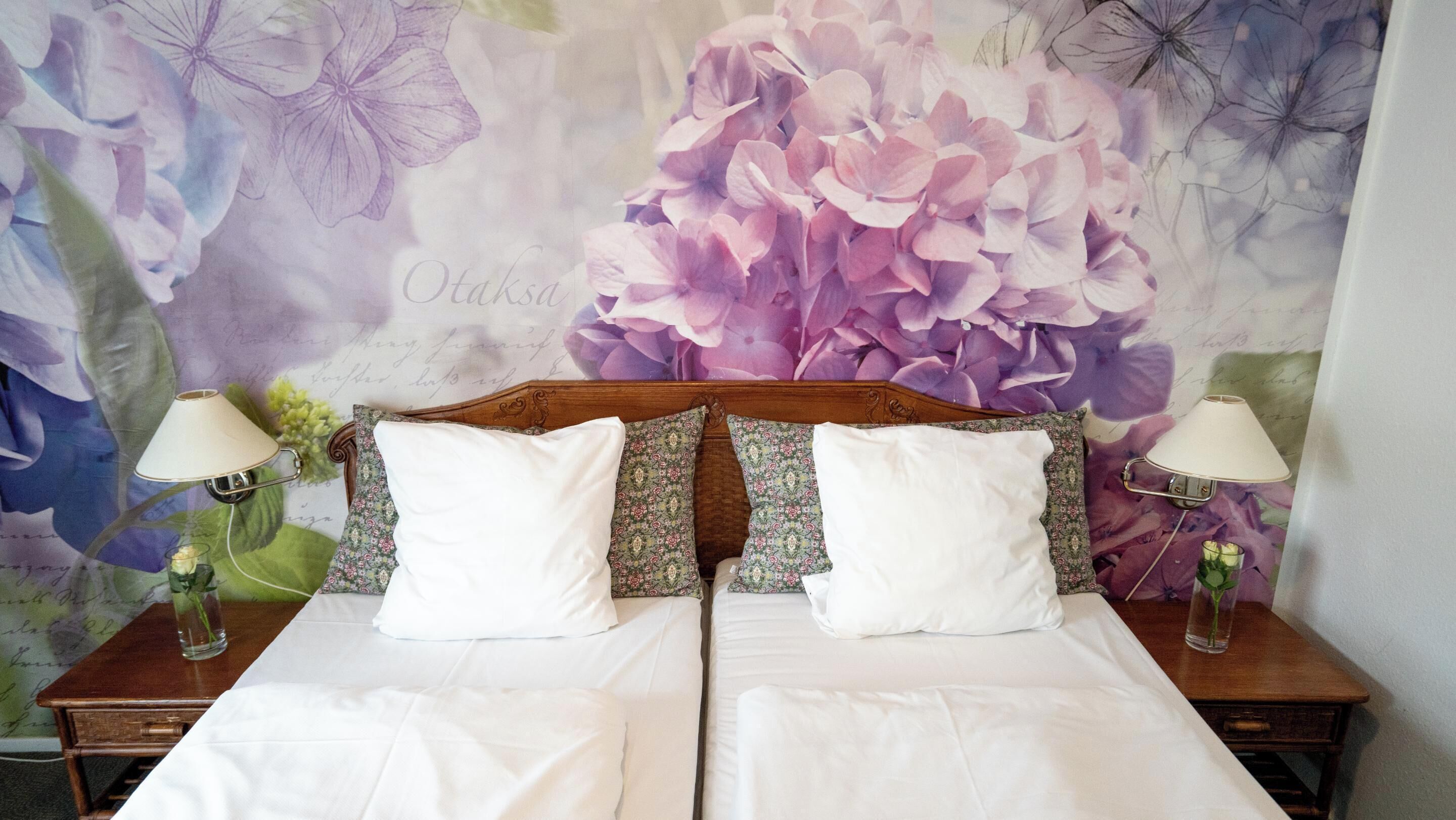 Una cama doble con sábanas blancas, dos mesitas de noche con lámparas y un fondo de papel tapiz con motivos florales.