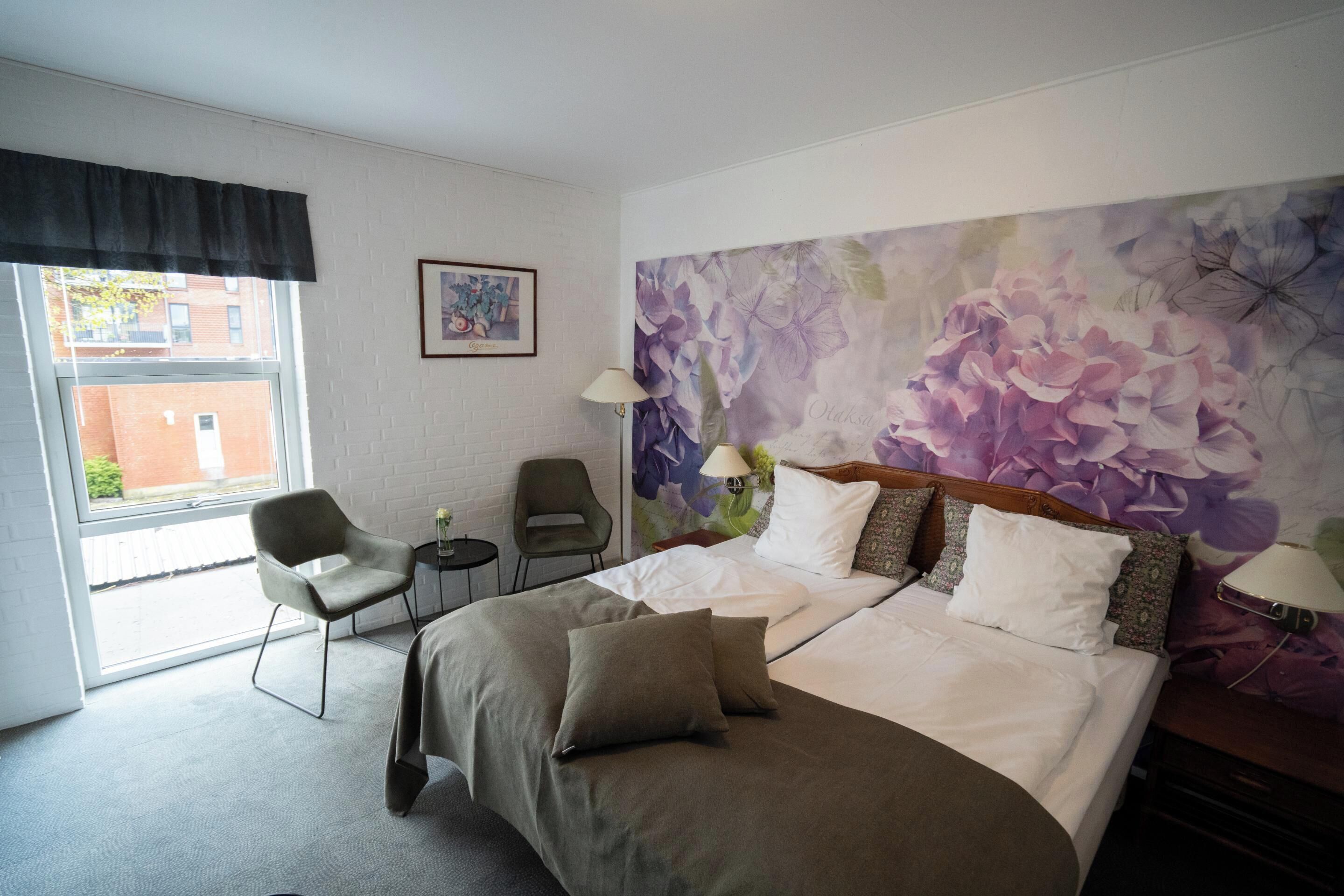 Habitación de hotel con una cama grande, dos sillas, una mesita y ventana con vistas a edificios.