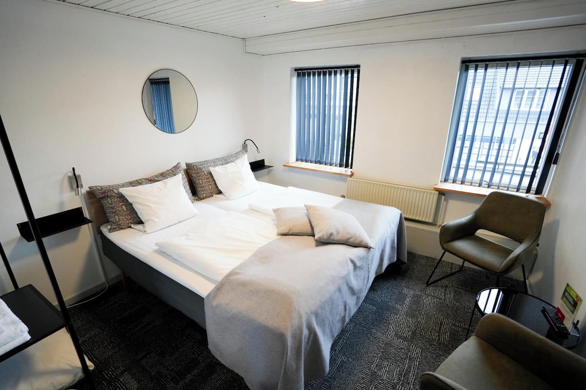 A hotel room with a bed, a chair, a desk, a mirror, and two windows with blinds.