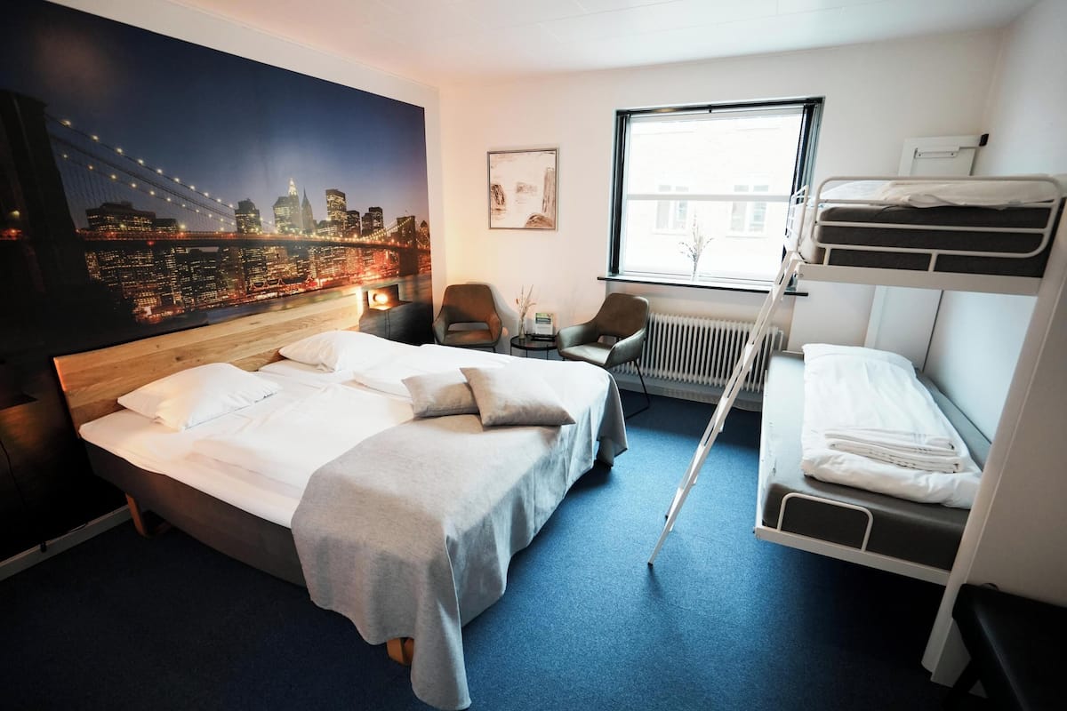 A hotel room with a large bed, a smaller bed, a desk, two chairs, a radiator, and a window with a cityscape view.