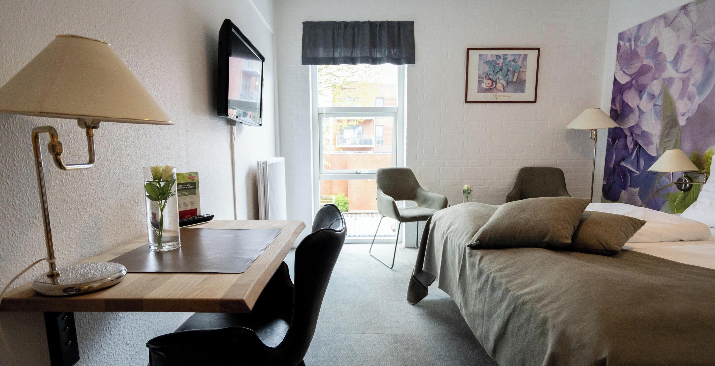 Ein Hotelzimmer mit Bett, Schreibtisch mit Lampe, Stuhl, Wandfernseher und Fenster mit Aussicht.