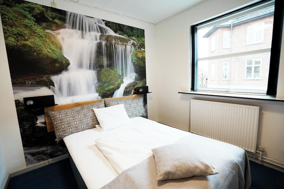 A hotel room with a bed, a mural of a waterfall, a window with a view of buildings, and a radiator.