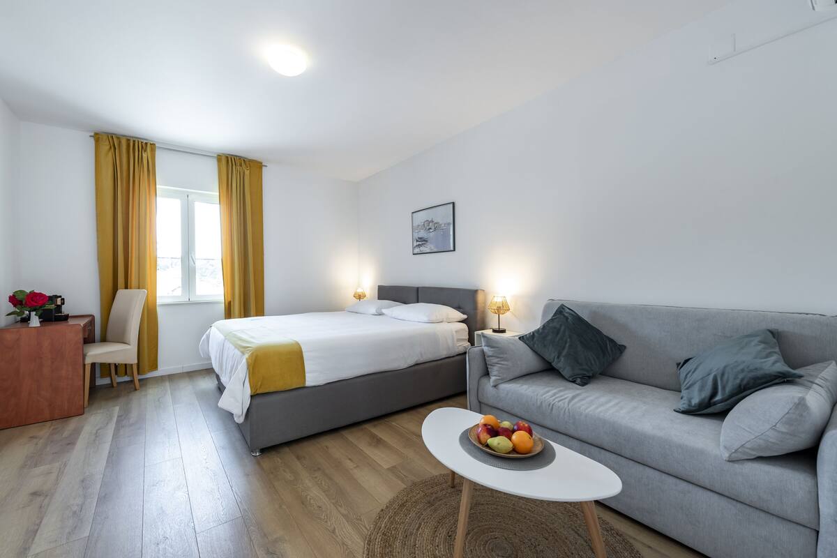 A modern hotel room with a bed, a grey sofa, a small table with fruit, a desk, and a chair.