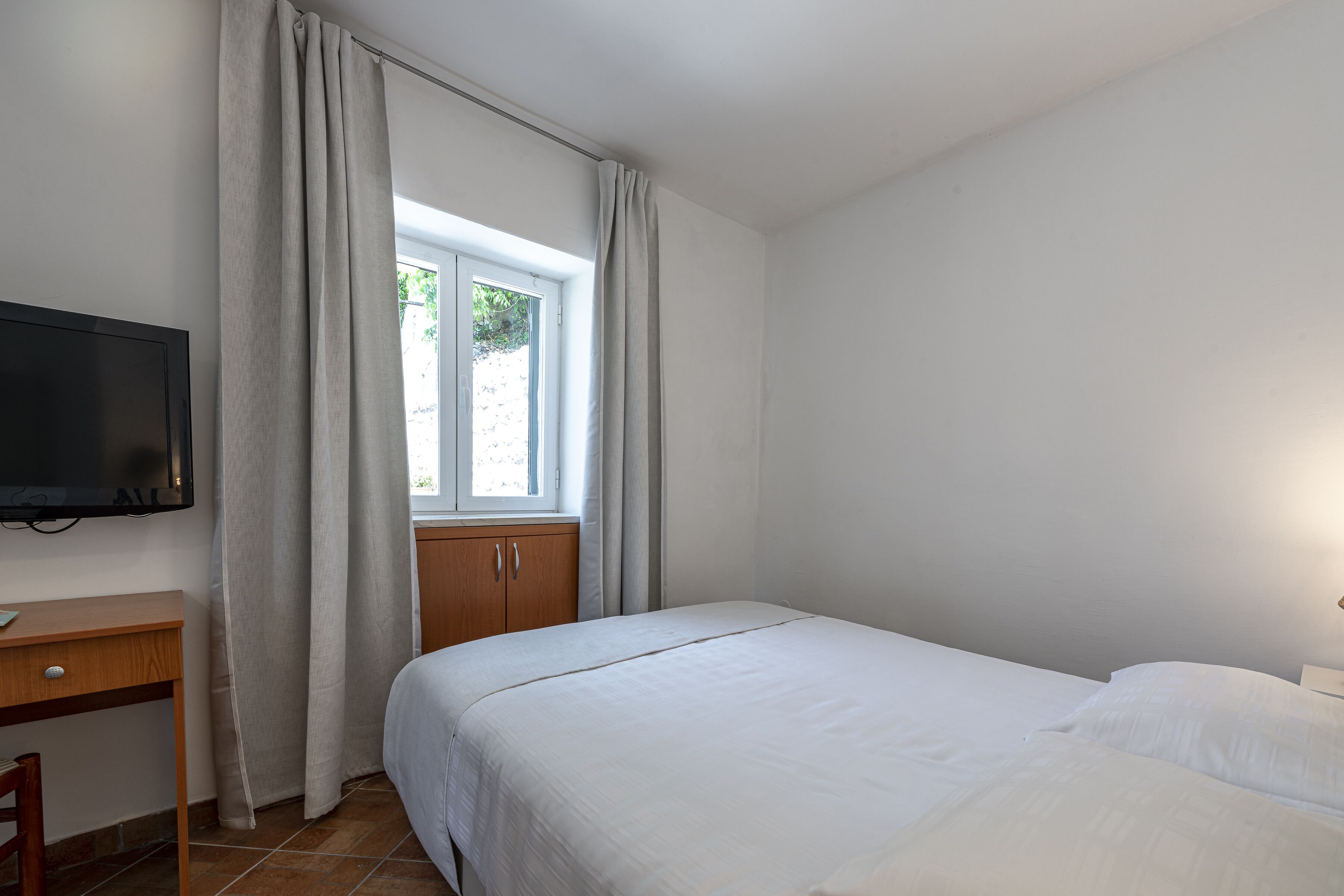 A bedroom with a bed, a desk, a TV, and a window with curtains.