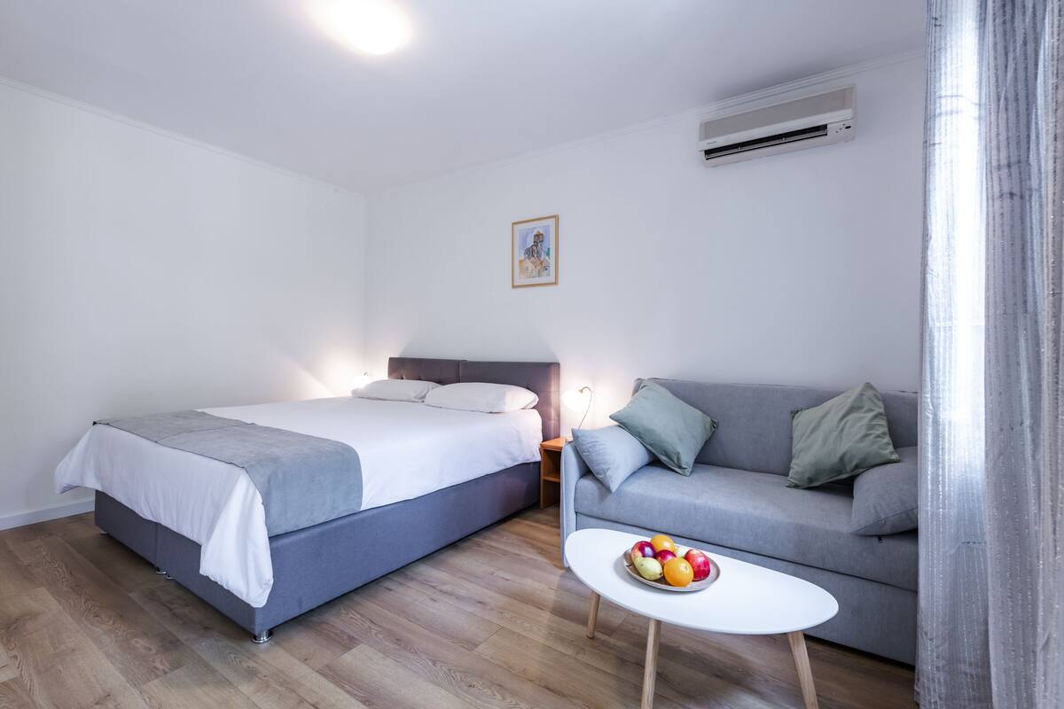A bedroom with a bed, a gray sofa, a small table with fruit, and an air conditioning unit.