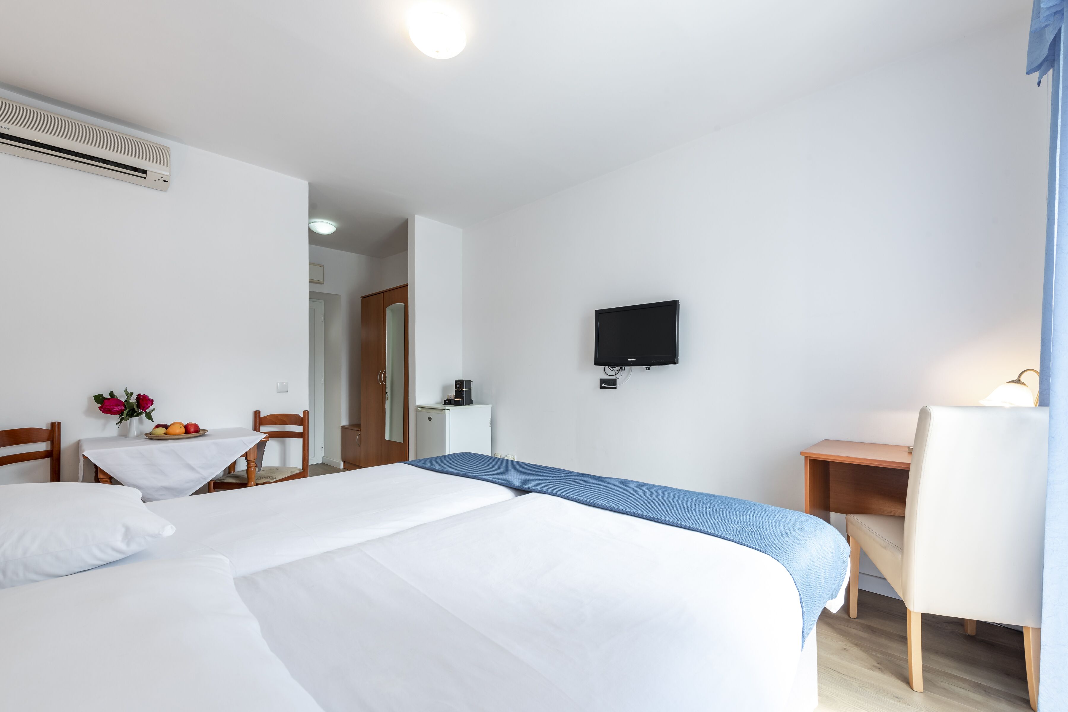 Hotellihuone, jossa on kaksi sänkyä, työpöytä, tuoli, televisio sekä pieni pöytä, jonka päällä on kukkia ja hedelmiä.