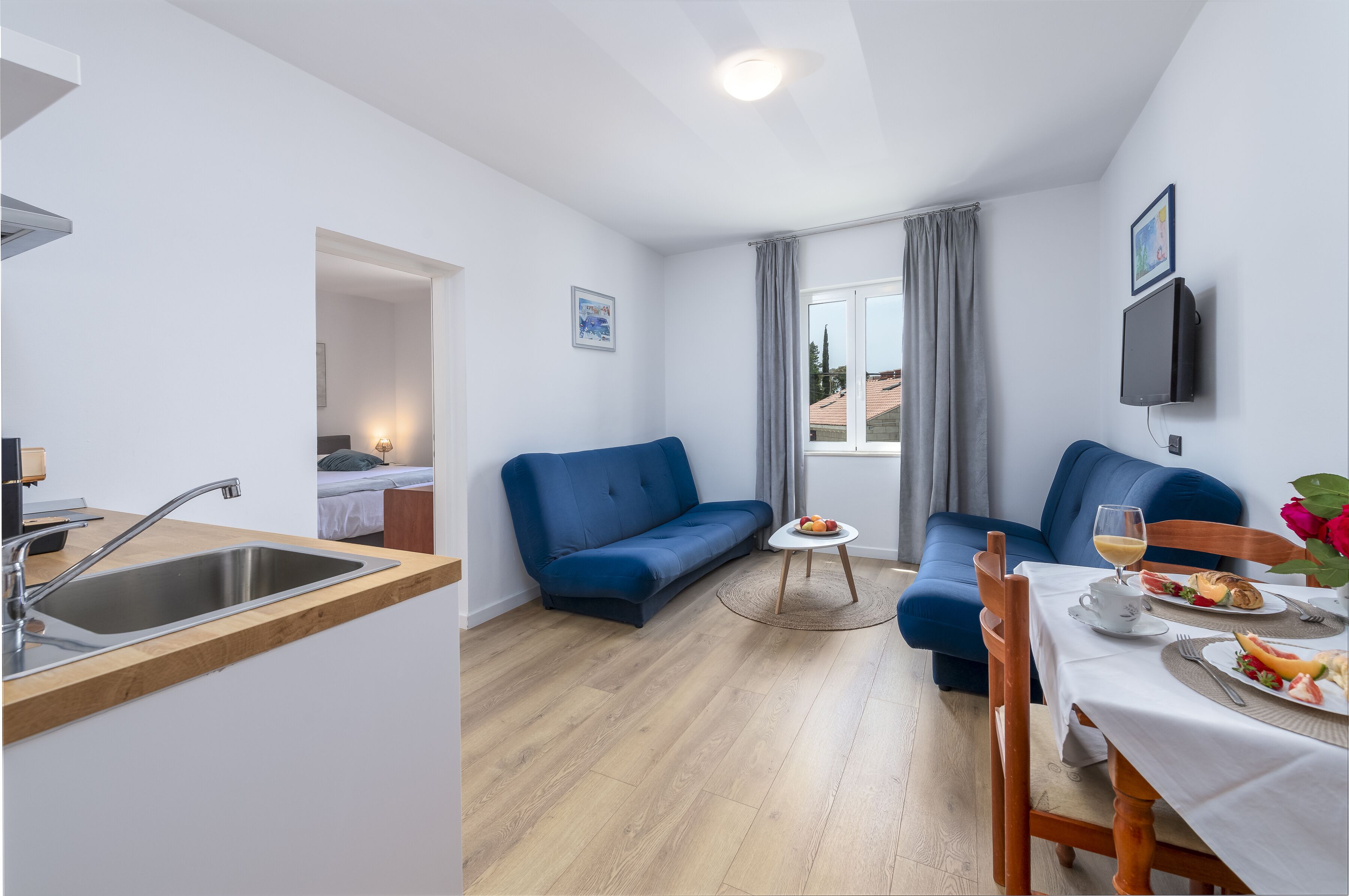 Ein modernes Wohnzimmer mit einem blauen Sofa, einem Esstisch für zwei Personen und einer Küchenzeile mit Spüle und einem fensterbepflanzten Fenster.