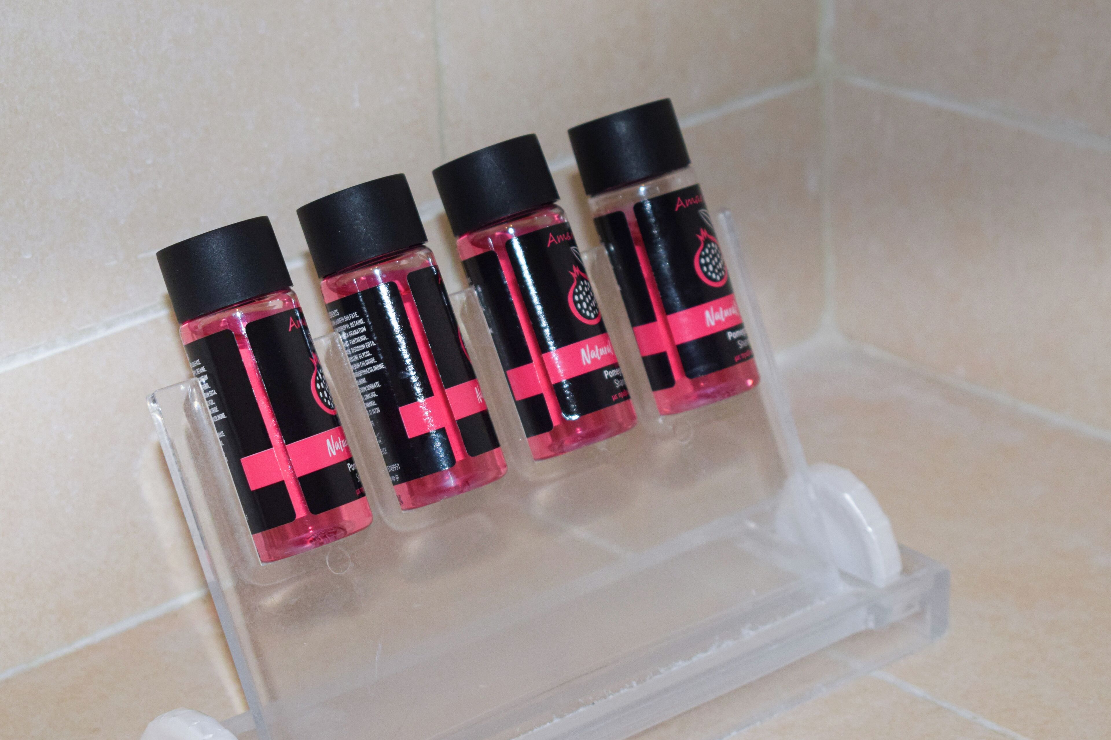 Un conjunto de cuatro botellas pequeñas con tapones negros y etiquetas rosas, dispuestas en un expositor de plástico transparente sobre una superficie de baldosas.
