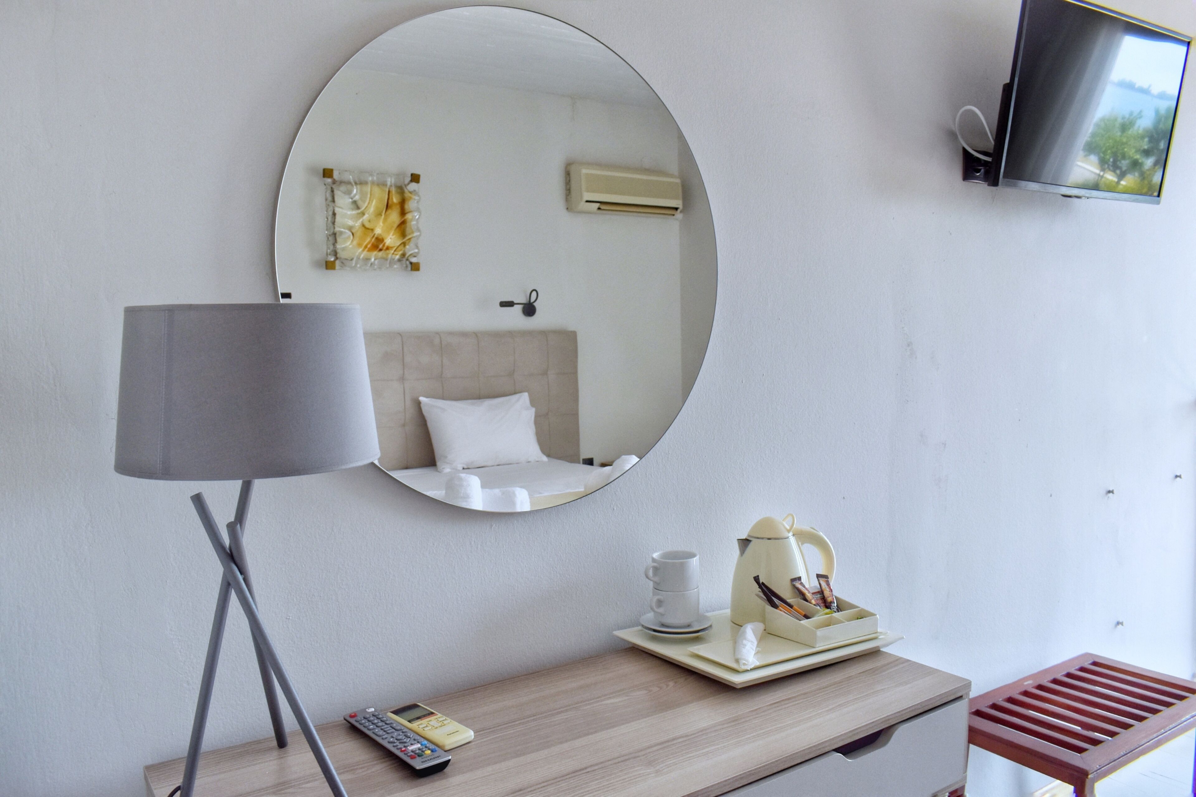 Een hotelkamer met een ronde spiegel, een nachtkastje met lamp, een aan de muur bevestigde televisie en een bed met hoofdeinde.