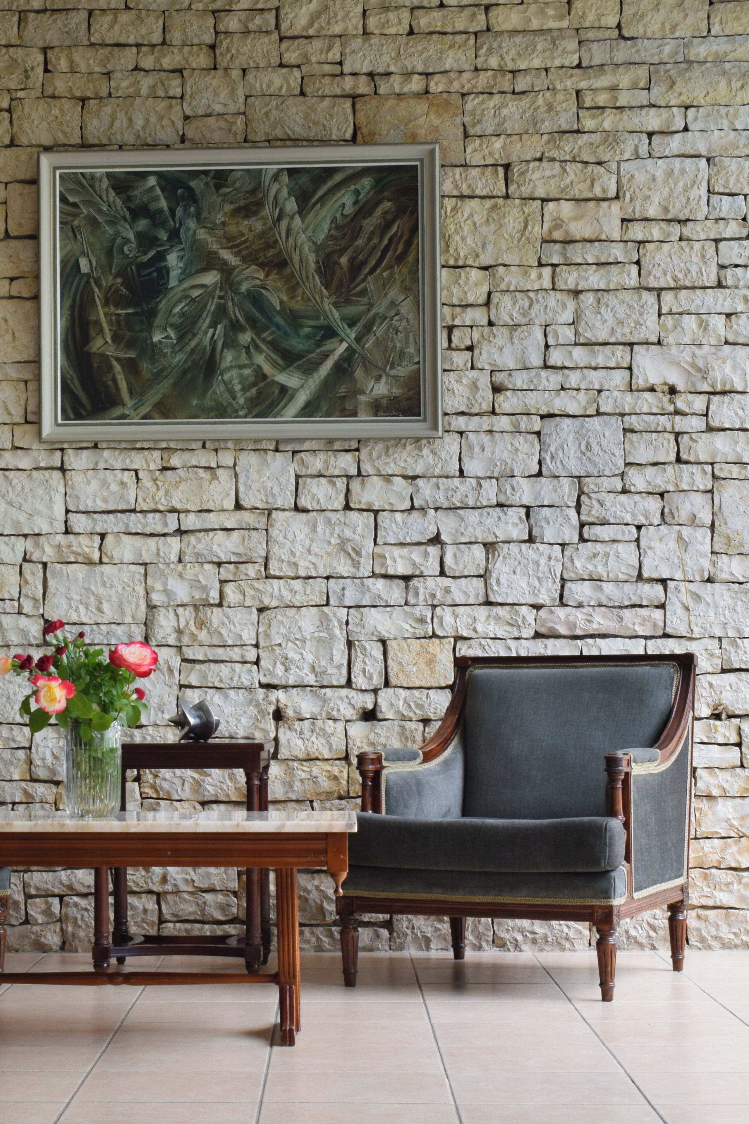 Un muro de piedra con un cuadro enmarcado, una mesa de madera con un jarrón de flores y una silla tapizada oscura.