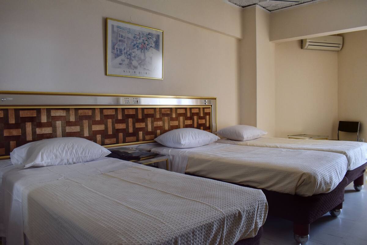 Ein Hotelzimmer mit zwei Betten, einem Kopfstück mit geometrischem Muster, einem gerahmten Bild an der Wand und einer Klimaanlage.