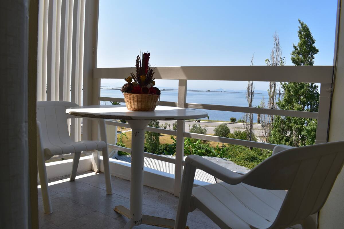 Ein Balkon mit Tisch und zwei Stühlen, mit Blick auf ein Gewässer und Grünflächen.