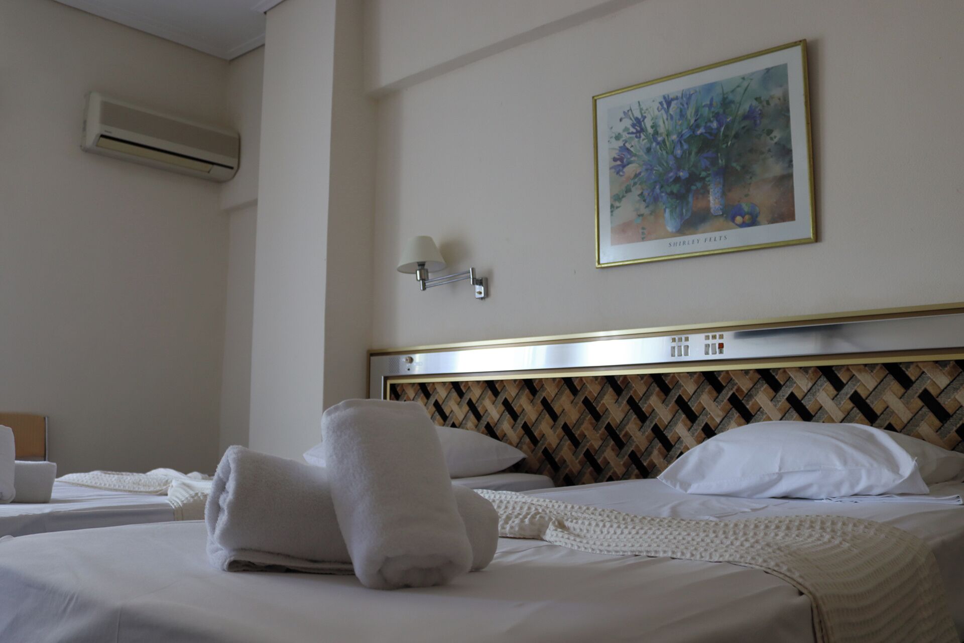 A hotel room with a bed, a wall-mounted air conditioner, a framed painting, and a bedside lamp.