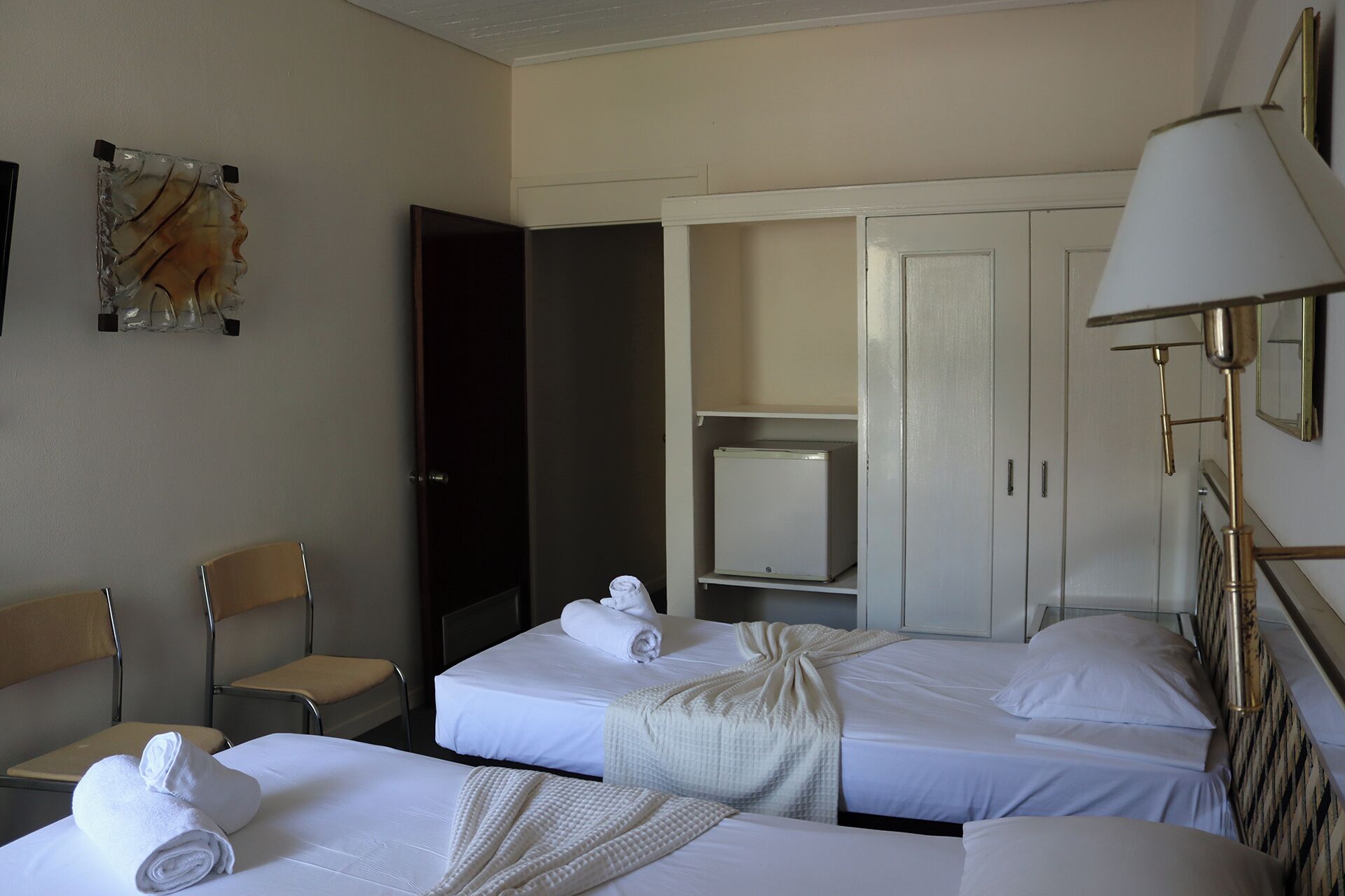 Habitación de hotel con dos camas, un escritorio, una silla, un armario y una lámpara.