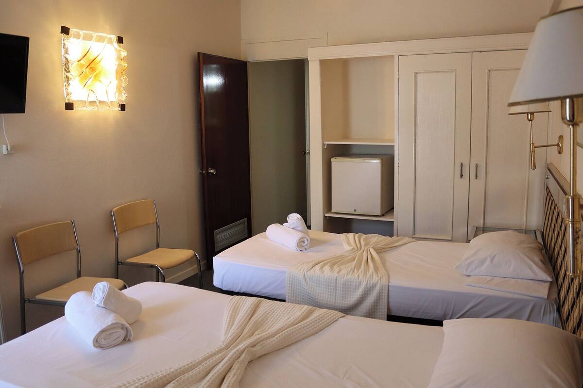 Ein Hotelzimmer mit zwei Einzelbetten, einem Schreibtisch, einem Stuhl, einem Kleiderschrank und einem kleinen Fernseher.