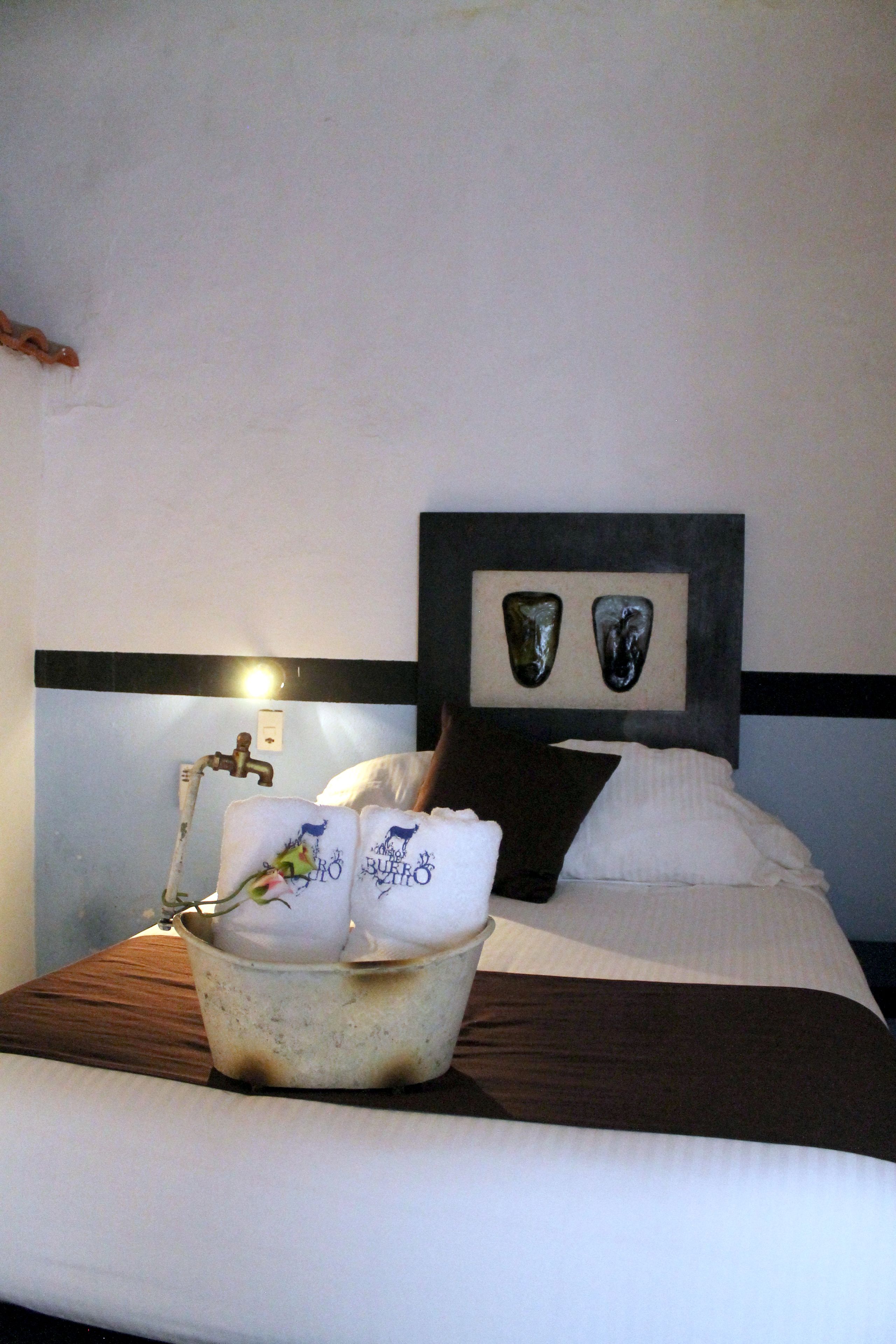 Habitación de hotel con cama, mesita de noche, un cubo con toallas y cabecero decorado con dos detalles.