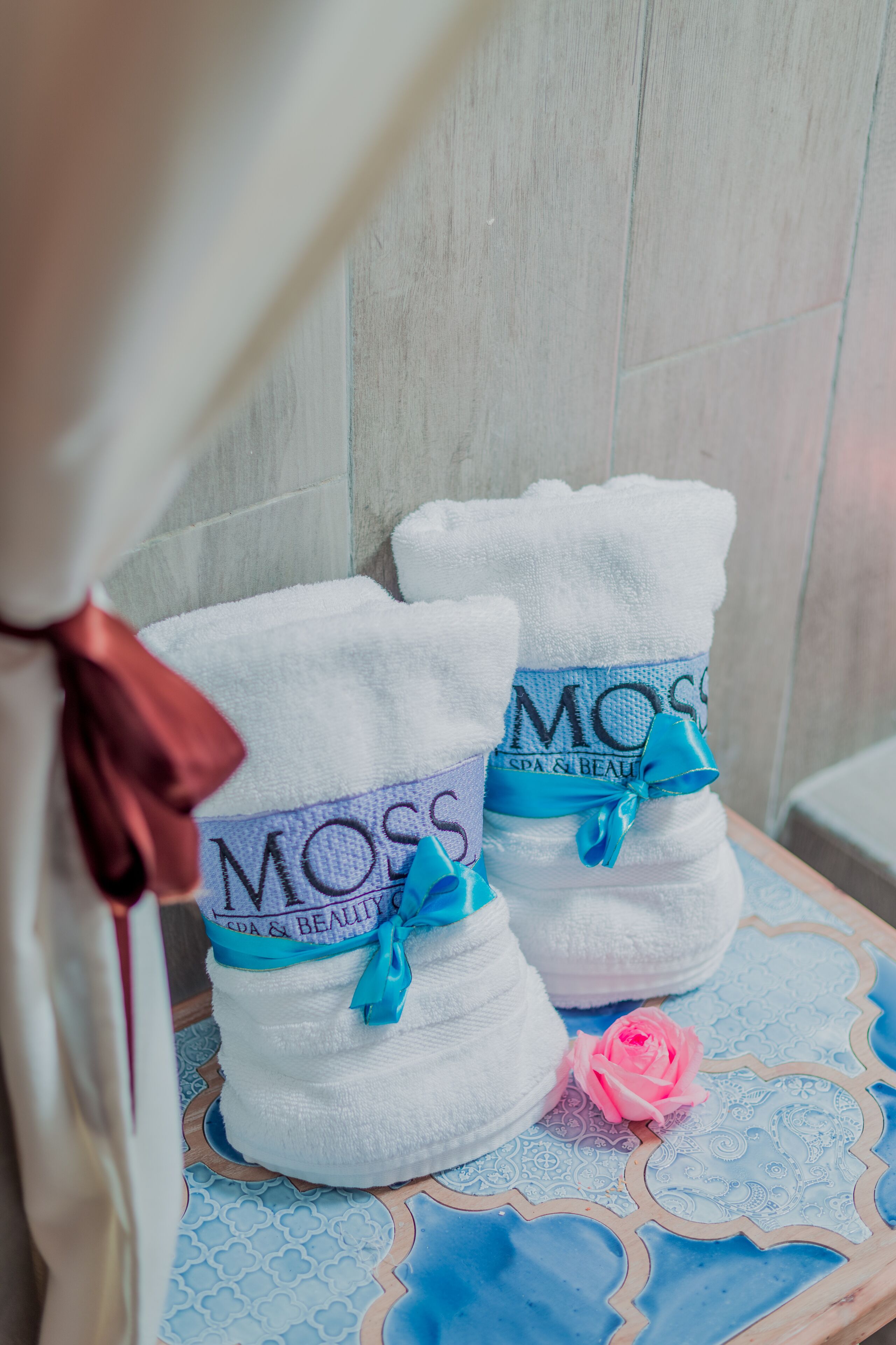Two white towels with "MOSS SPA & BEAUTY" text and a blue ribbon, placed on a patterned tablecloth.