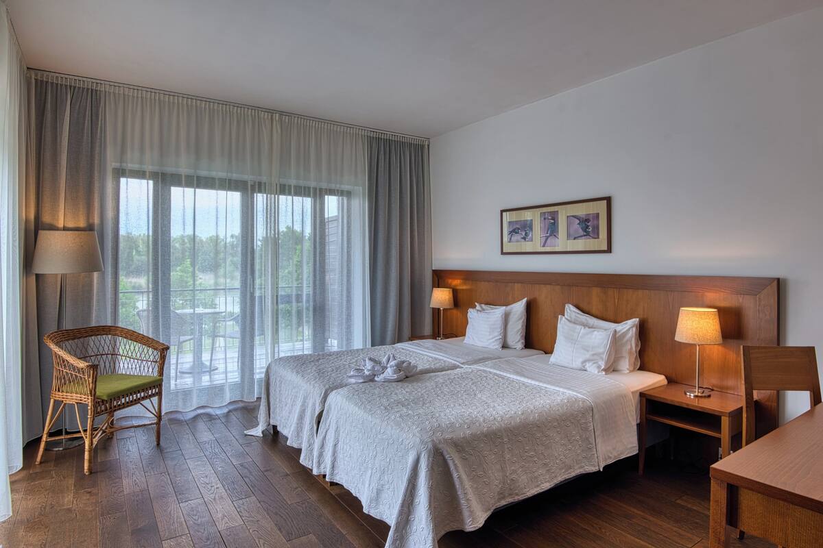 Een hotelkamer met een groot bed, een houten hoofdbord, een houten bureau, een rieten stoel en een groot raam met gordijnen.