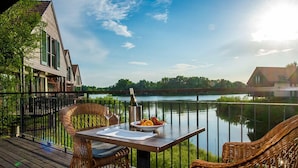 A wooden deck with wicker chairs and a table set for two, overlooking a calm lake and a distant tree line. - Tisza Balneum Thermal Hotel (Tiszafured)