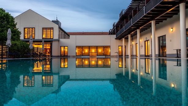 A modern hotel with a clear swimming pool, multiple rooms with warm lighting, and a covered outdoor area with umbrellas. - Tisza Balneum Thermal Hotel (Tiszafured)
