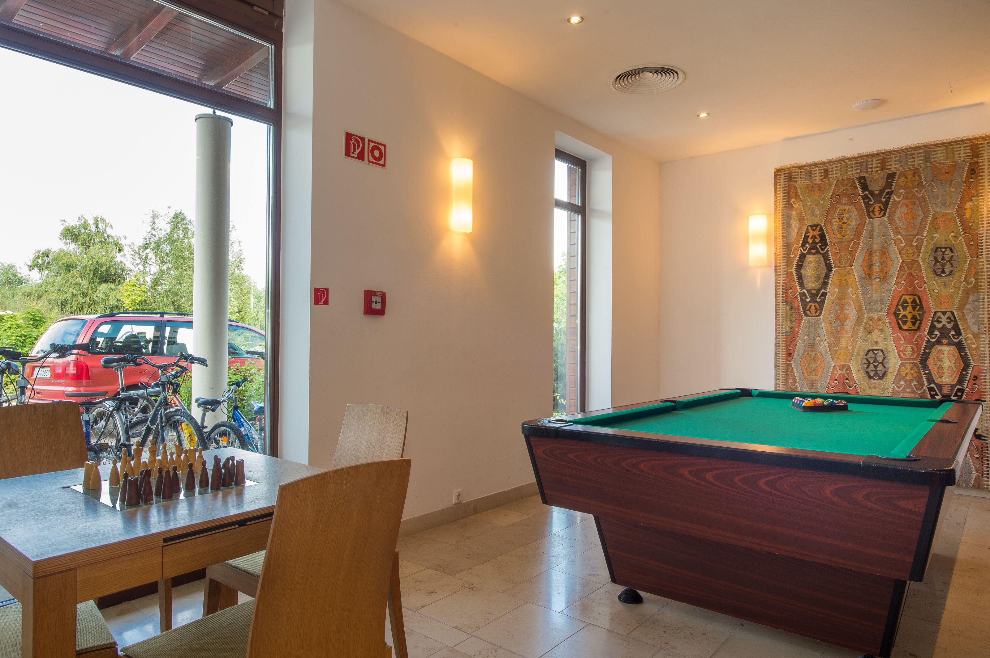 Ein Billardtisch, Stühle und ein Schachbrett in einem Raum mit einem großen Fenster, einem gemusterten Wanddesign und einem Feuerschutzschild.
