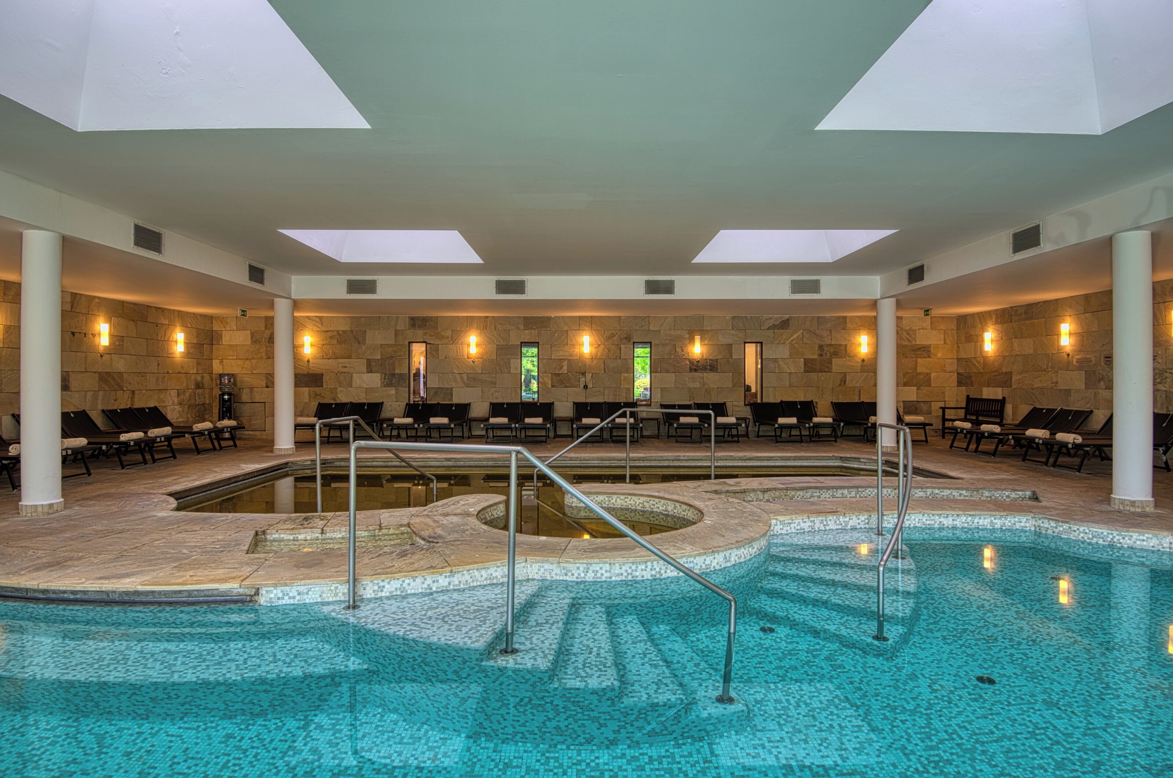 Piscina interior moderna com um elemento central de água, rodeada por espreguiçadeiras e paredes de pedra.
