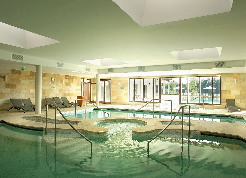Una piscina interior con zonas iluminadas por el sol, sillas de descanso y vistas al exterior.