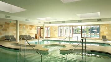 Una piscina interior con zonas iluminadas por el sol, sillas de descanso y vistas al exterior.