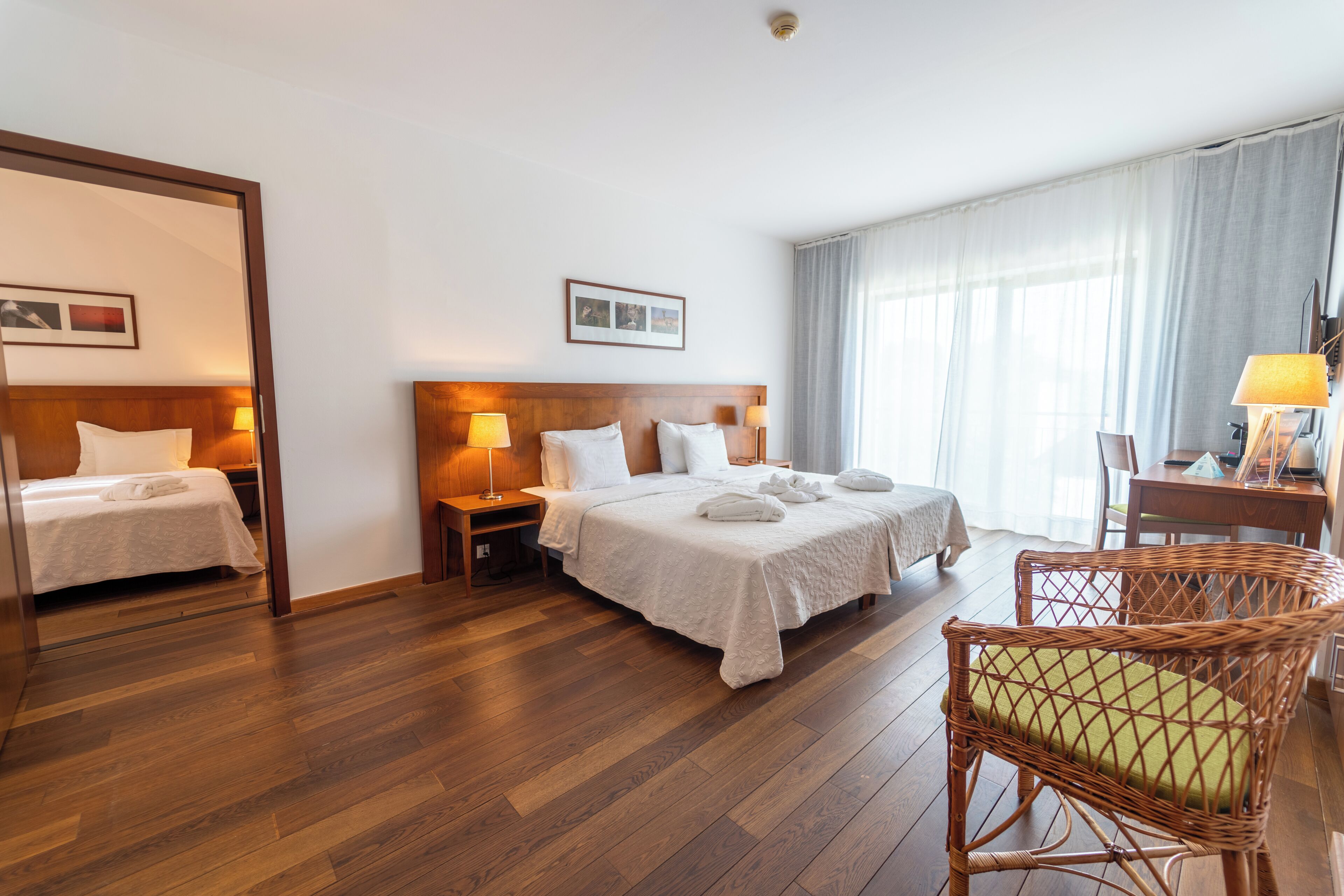 A hotel room with two beds, a wooden floor, a wicker chair, and a desk with a lamp.