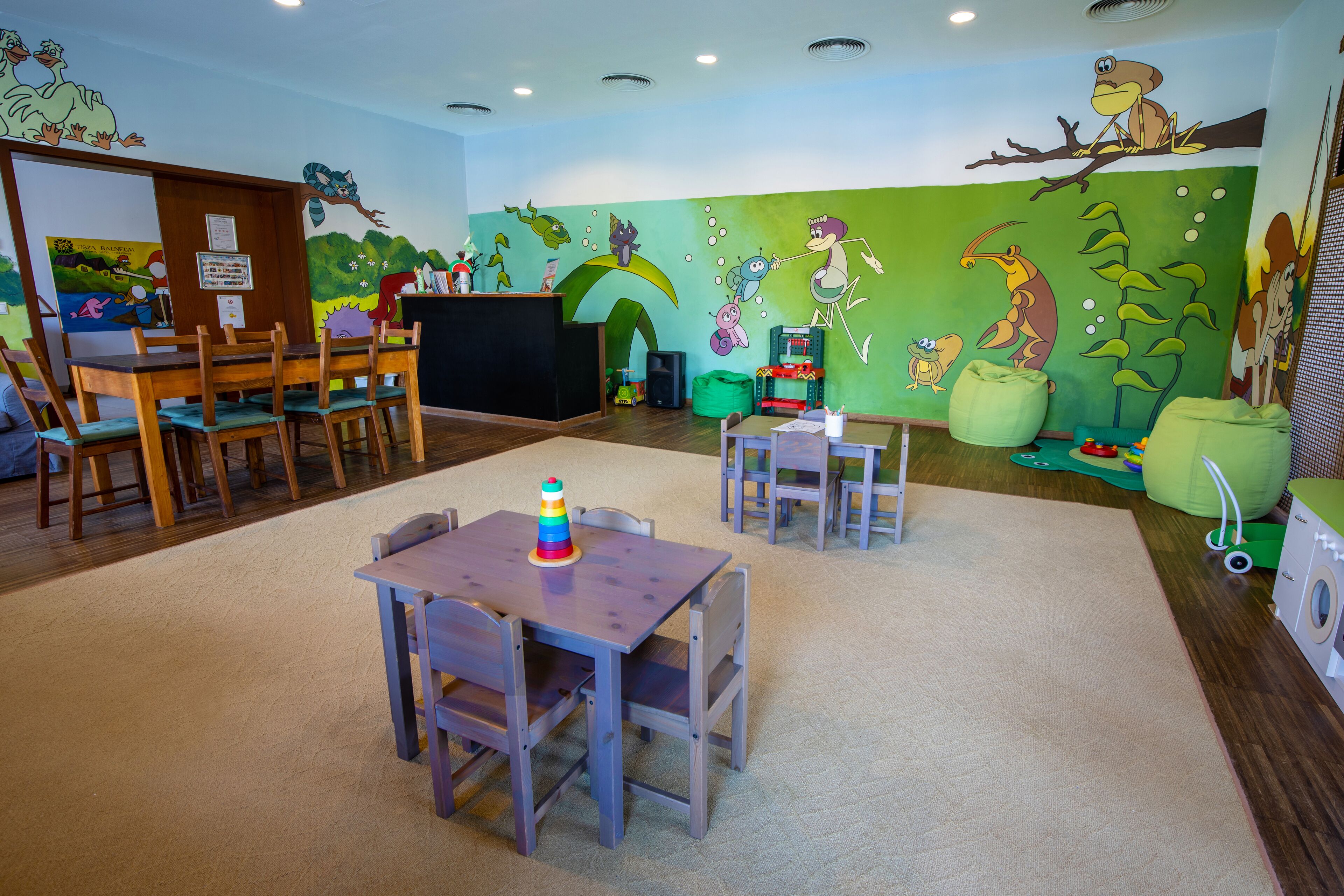 Een kinderspeelruimte met een muurschildering, houten tafels en stoelen.