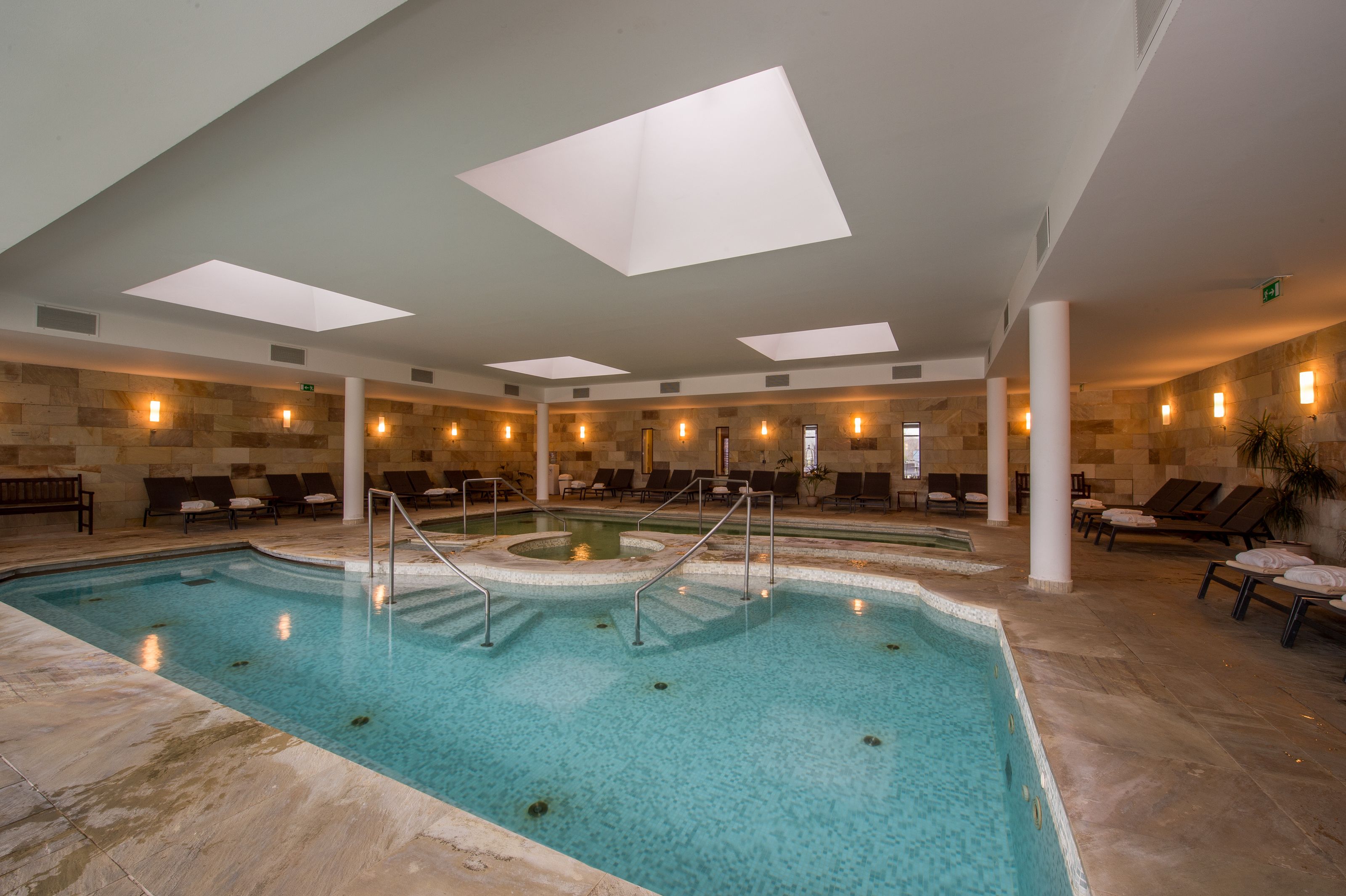 Une piscine intérieure à l’eau claire, entourée de fauteuils et de chaises longues.