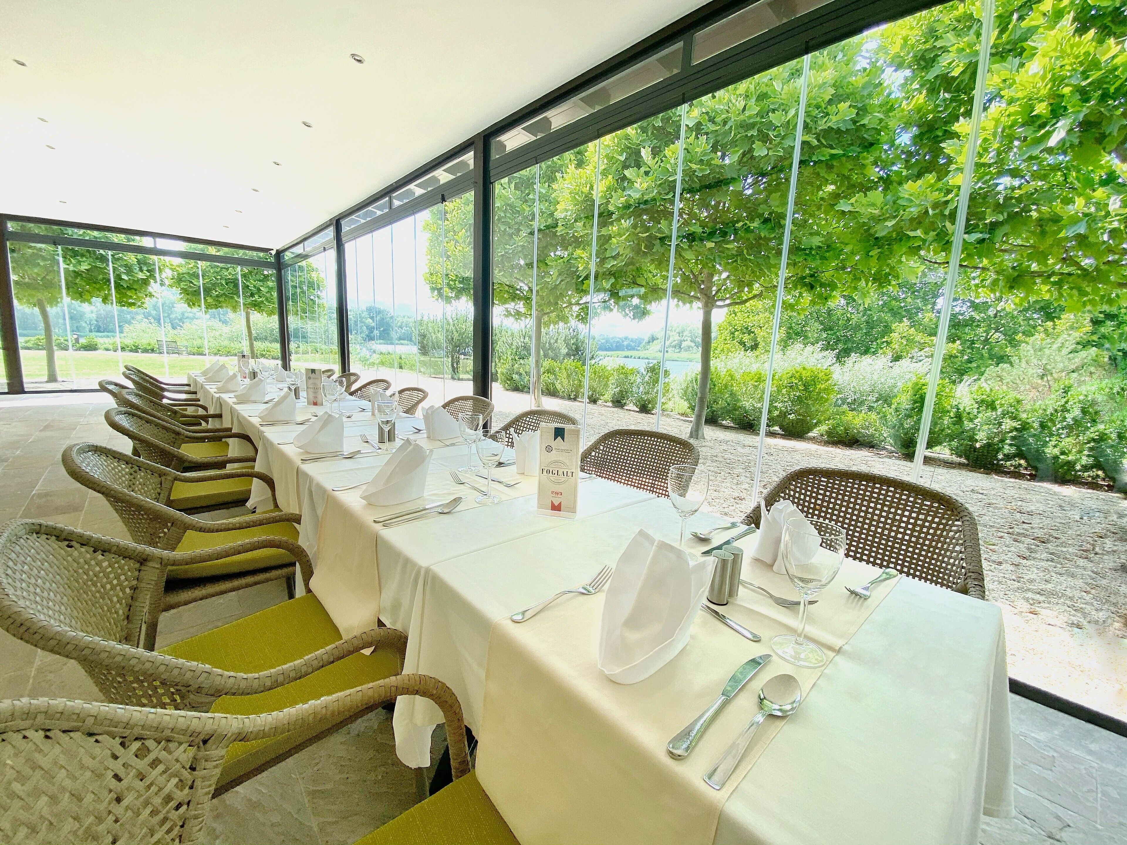 Una larga mesa de comedor preparada para una comida, con sillas de mimbre, manteles blancos y vistas al exterior.